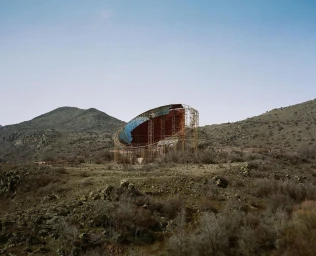 Large rusty industrial structure surrounded by scaffolding in a dry mountainous landscape under a pale sky