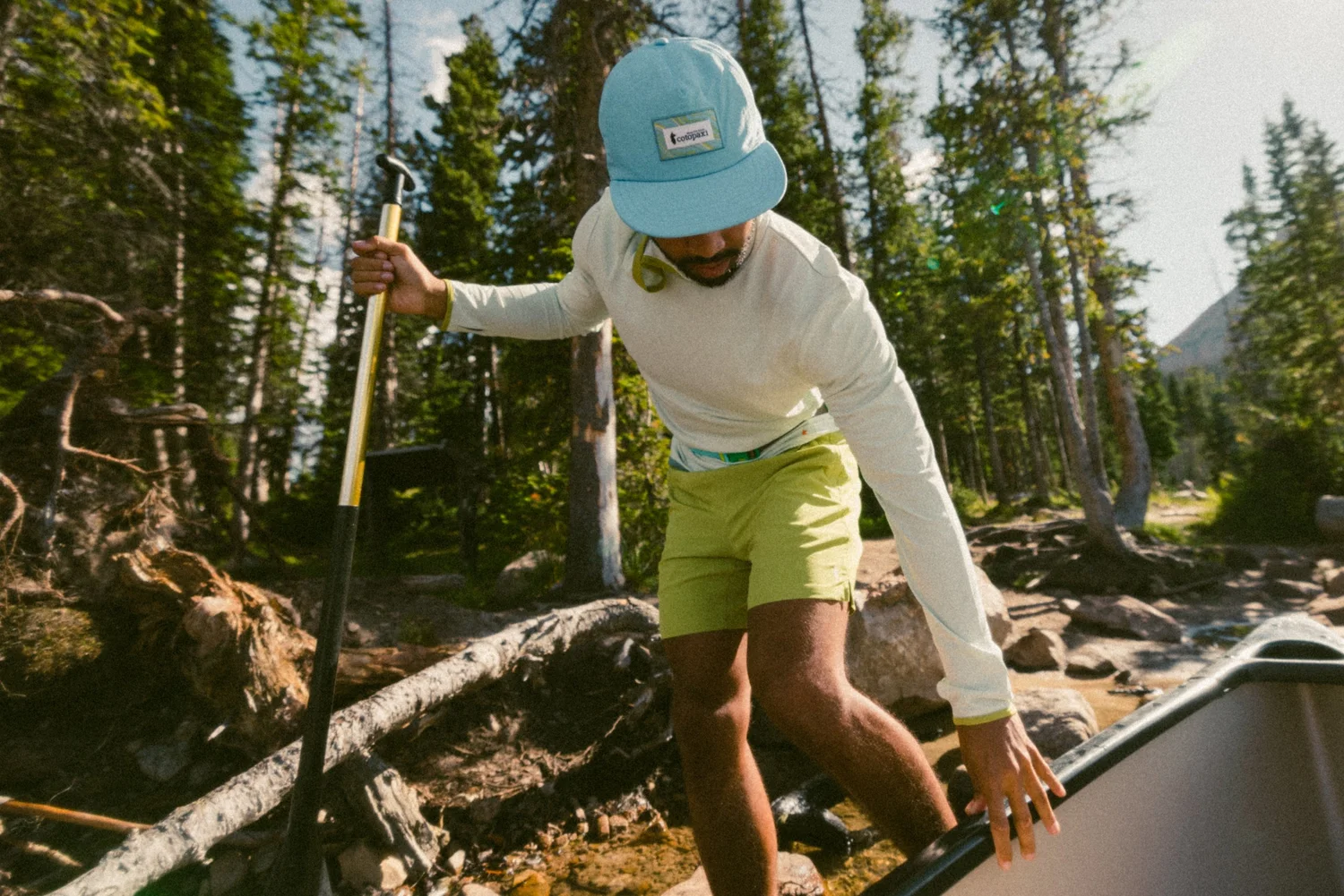 A man wades across a shallow stream next to a canoe, wearing a light blue cap, long-sleeved shirt, and green shorts in a lush forest.