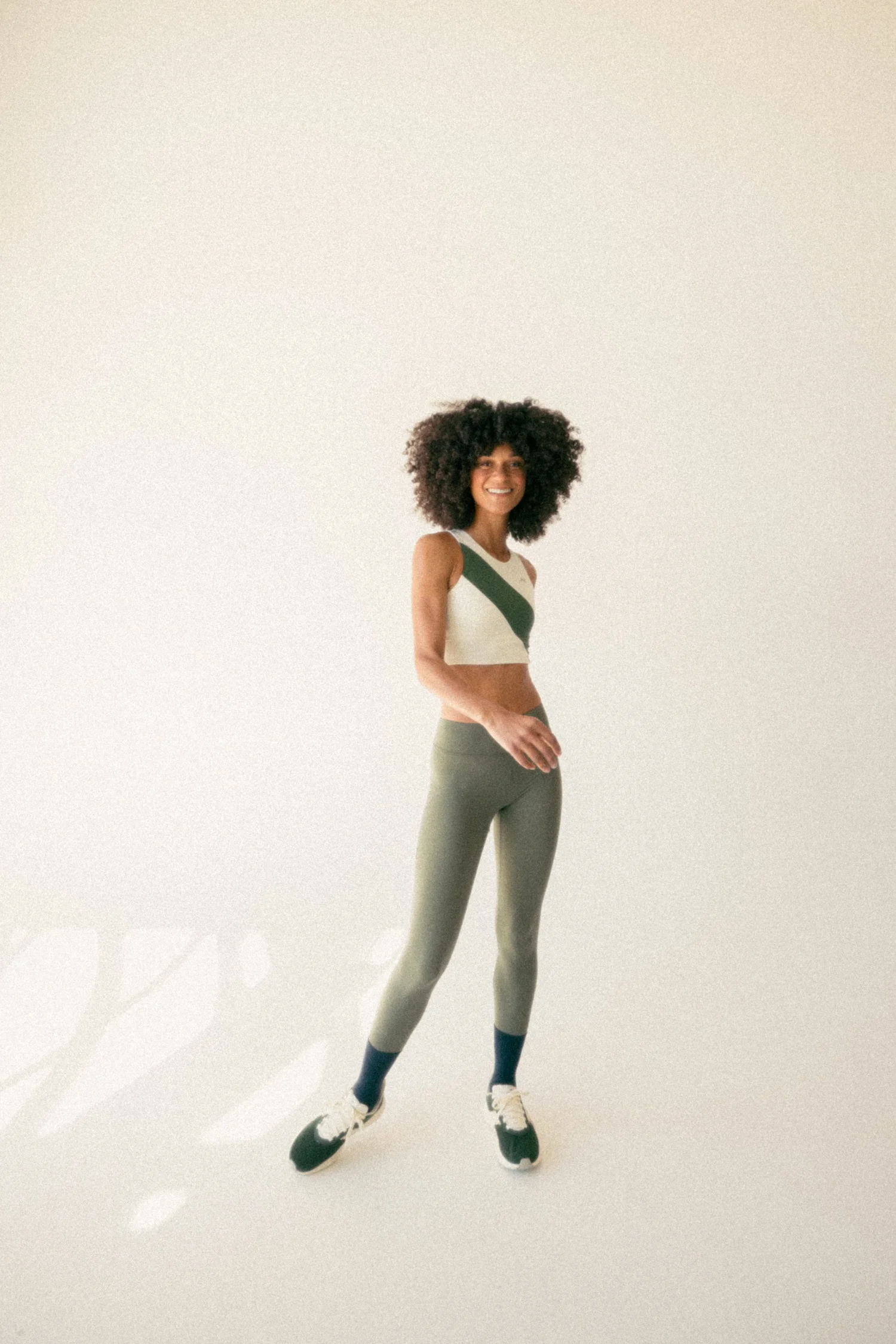 Athletic woman in a two-piece activewear outfit with green leggings and white crop top, standing in a brightly lit studio setting.