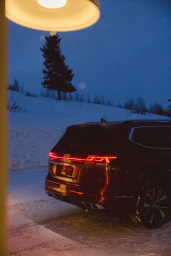Rear three-quarter view of a dark red SUV parked in snow at dusk with glowing red taillights and an overhead warm light.