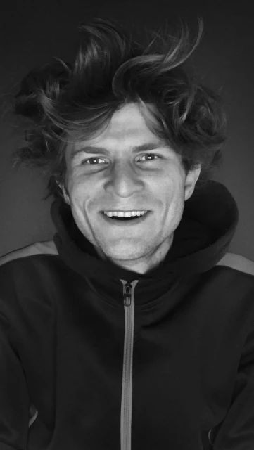 Portrait of a smiling adult male with messy brown hair wearing a zipped-up hoodie against a dark grey background in black and white.