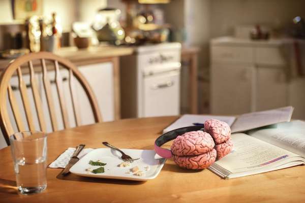 Two brain-shaped objects on an open book next to headphones and a plate with food remnants, and a glass of water on a wooden table.