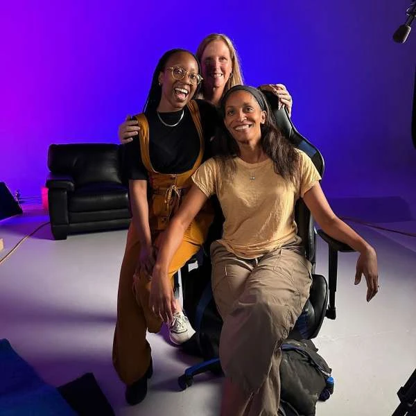Three women pose together indoors, one seated in a gaming chair, smiling. Purple and blue lighting creates a vibrant backdrop in the photo studio.