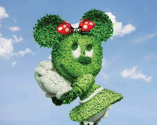 Minnie Mouse topiary with green foliage, red ladybug bow, and white gloves stands outdoors against blue sky with white clouds.