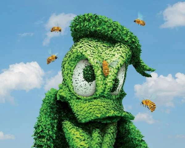 Green topiary art of Donald Duck with bees buzzing around it against a blue sky with clouds, creating whimsical outdoor scene.