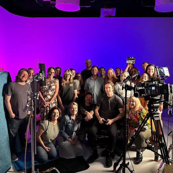 Group of people on a film set in front of purple and blue screen. Camera and lighting equipment are set up for the shoot.