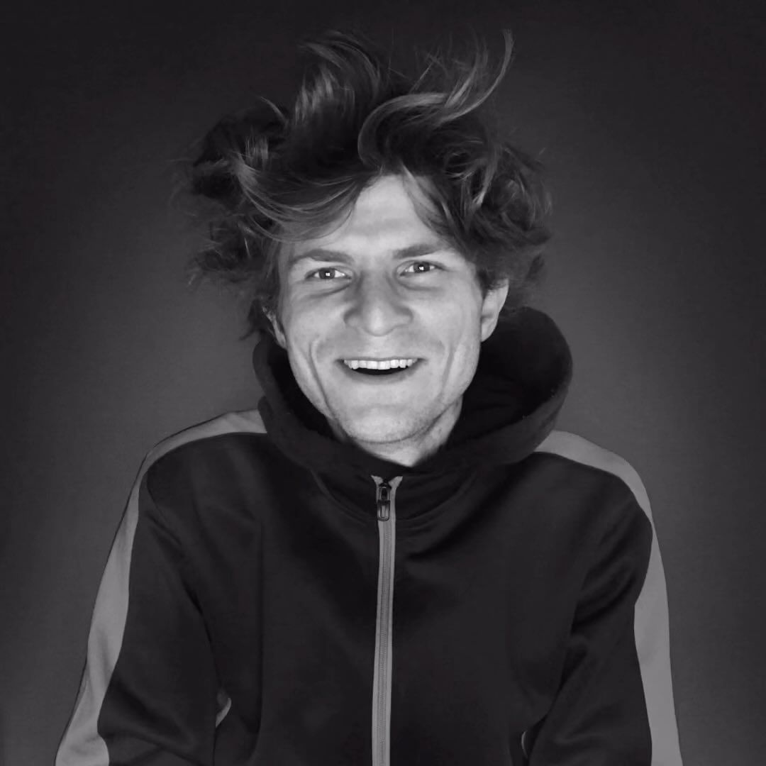 Black and white portrait of smiling adult male with tousled hair, wearing hooded sweatshirt, against a dark backdrop in a medium shot.