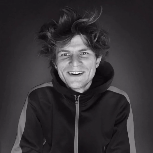 Black and white portrait of smiling adult male with tousled hair, wearing hooded sweatshirt, against a dark backdrop in a medium shot.