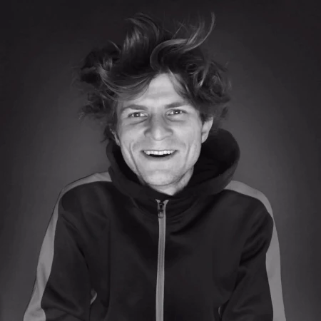 Black and white portrait of smiling adult male with tousled hair, wearing hooded sweatshirt, against a dark backdrop in a medium shot.