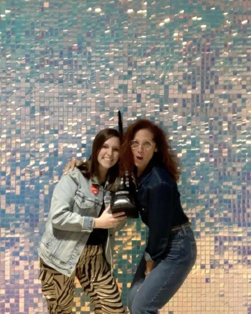 Two women pose indoors against a shimmering, square-tiled background. One woman has red hair and glasses. The other has zebra-striped pants.