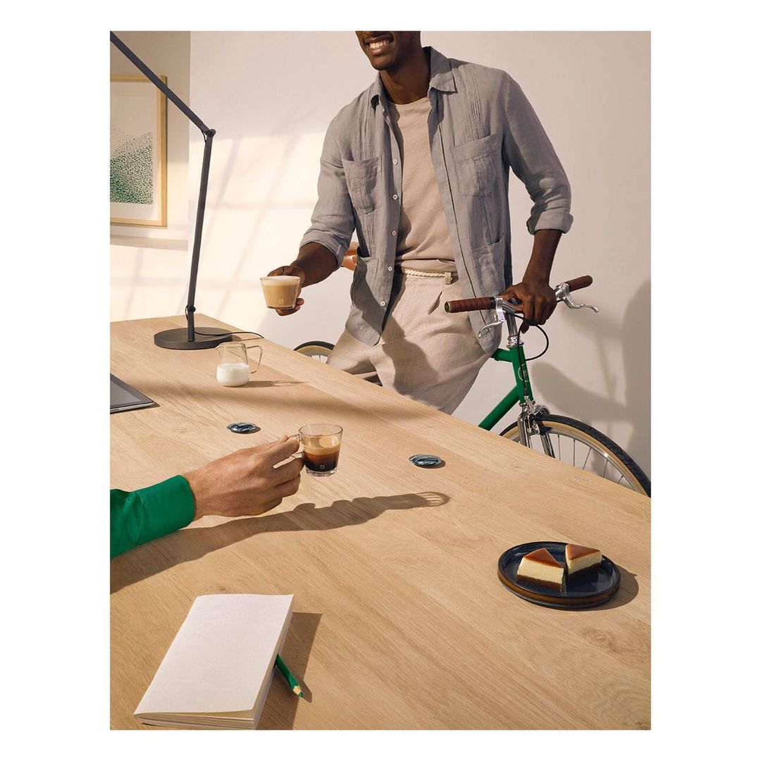 Two adults at a light wood table with a bicycle, drinks, dessert, notebook, and lamp, set indoors with soft, natural lighting.