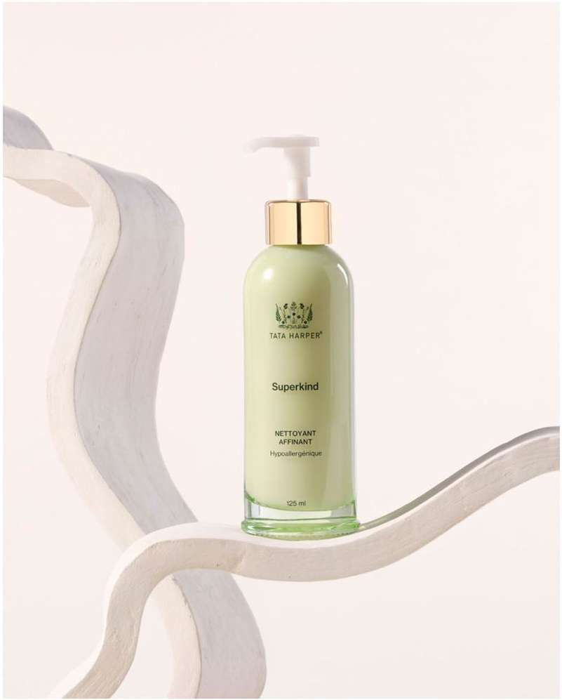 Tata Harper Superkind cleanser bottle in light green and gold, resting on a modern white sculpture against an off-white backdrop.