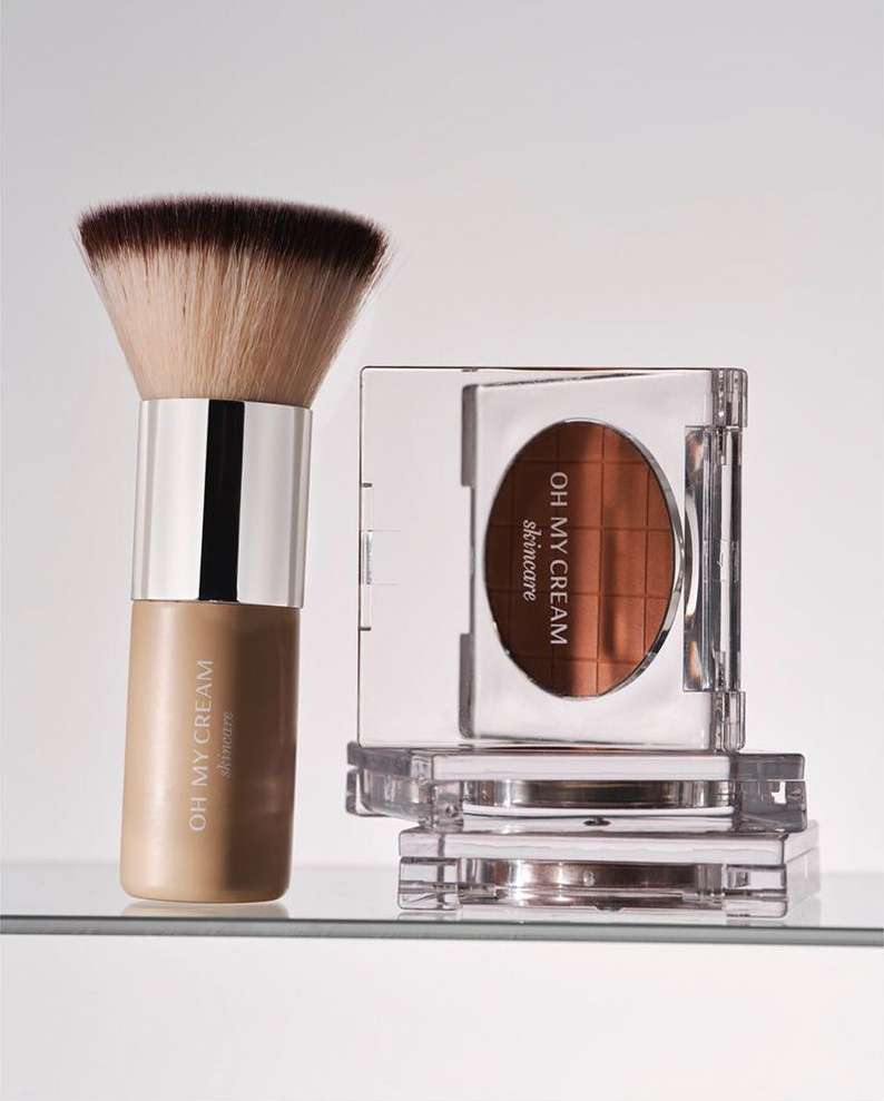 Cosmetic products on a shelf. A makeup brush and square container of bronzer skincare by OH MY CREAM are centered in the shot.