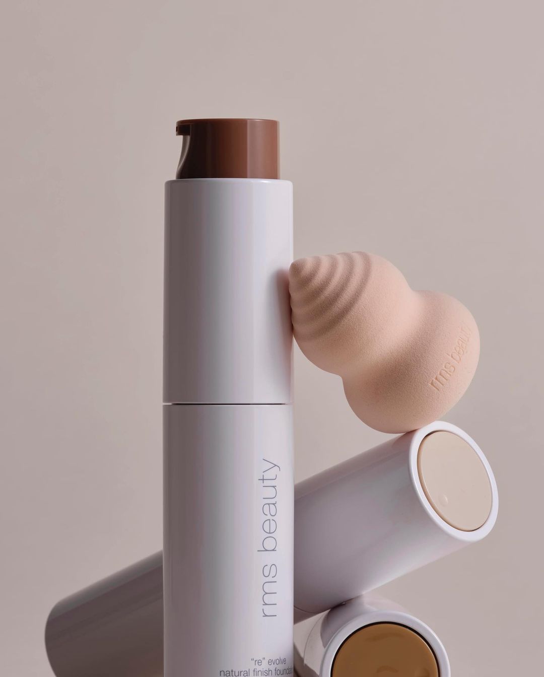 Close-up studio shot of RMS Beauty foundation bottle with a tan lid, cosmetic sponge and light neutral background for beauty advertising.