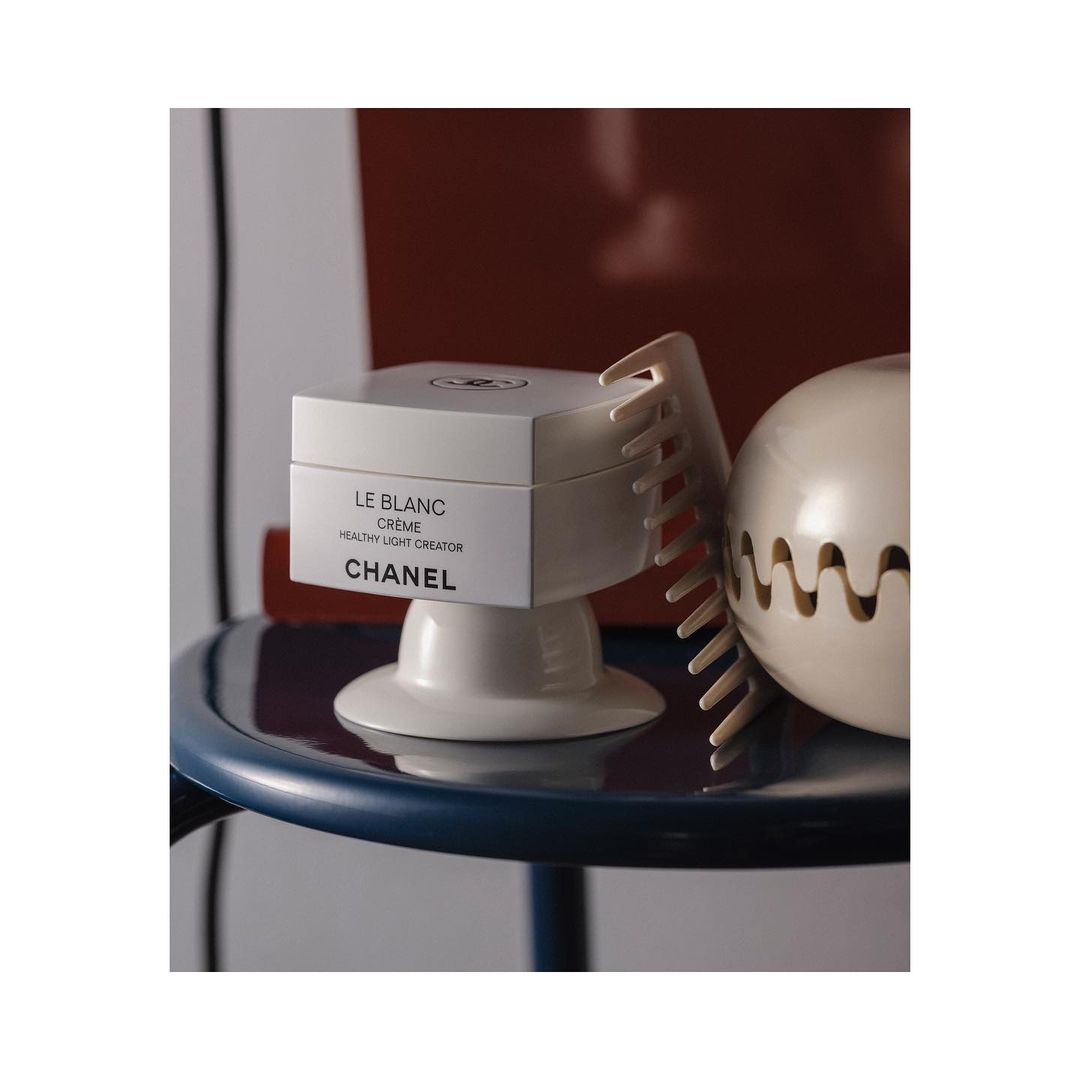 Still life shot of Chanel Le Blanc cream product, decorative hair comb, and a spherical sculpture on a blue side table indoors.