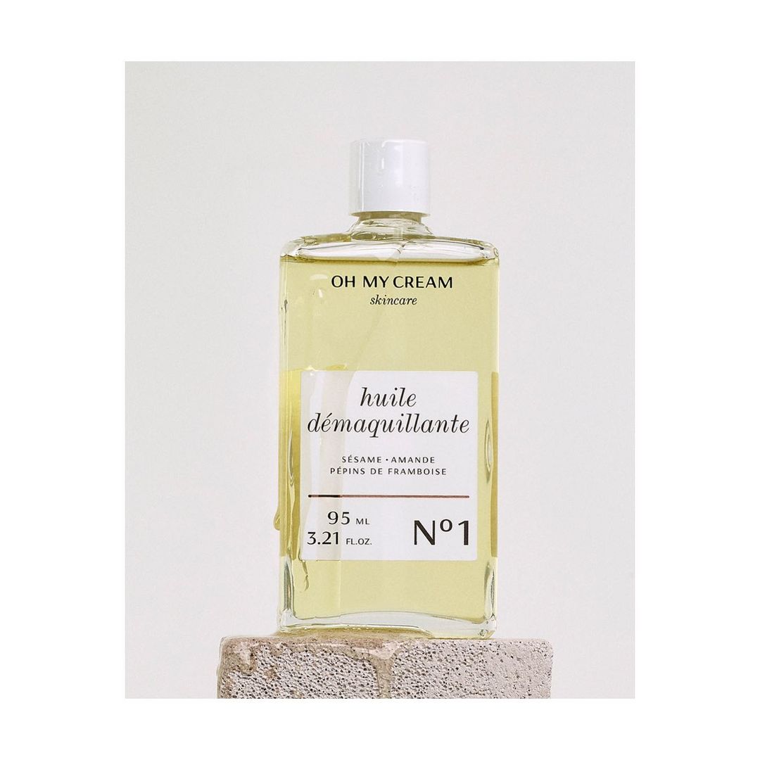 Square glass bottle of Oh My Cream skincare huile demaquillante resting on a stone block against a white background in a studio shot.