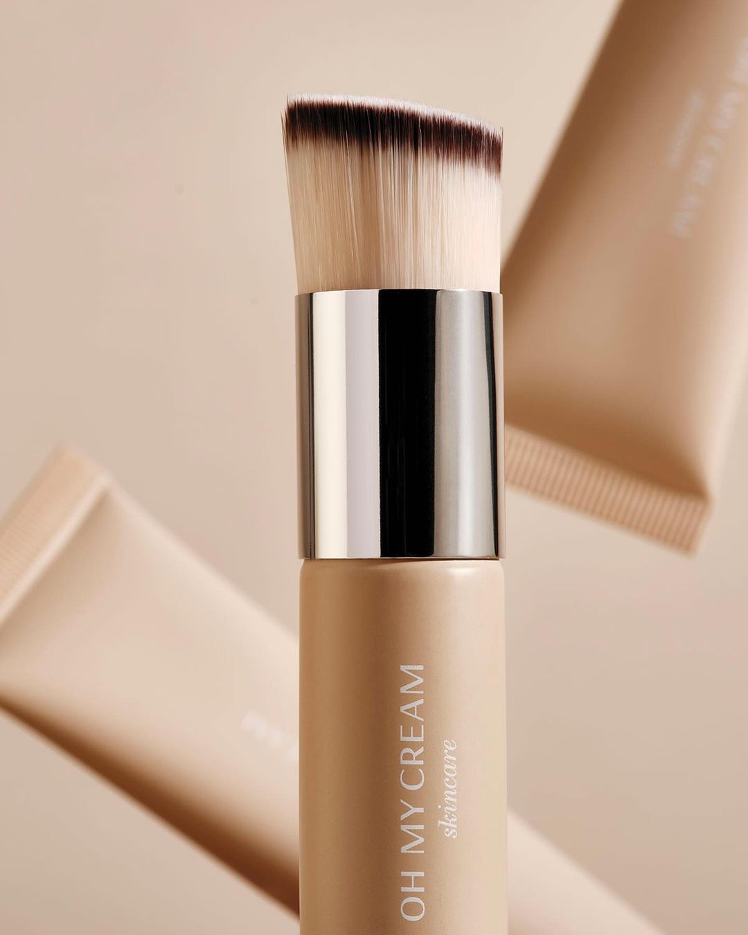 Beige background shot of a foundation brush with a metallic silver band and tube of skincare with text 'OH MY CREAM skincare'.
