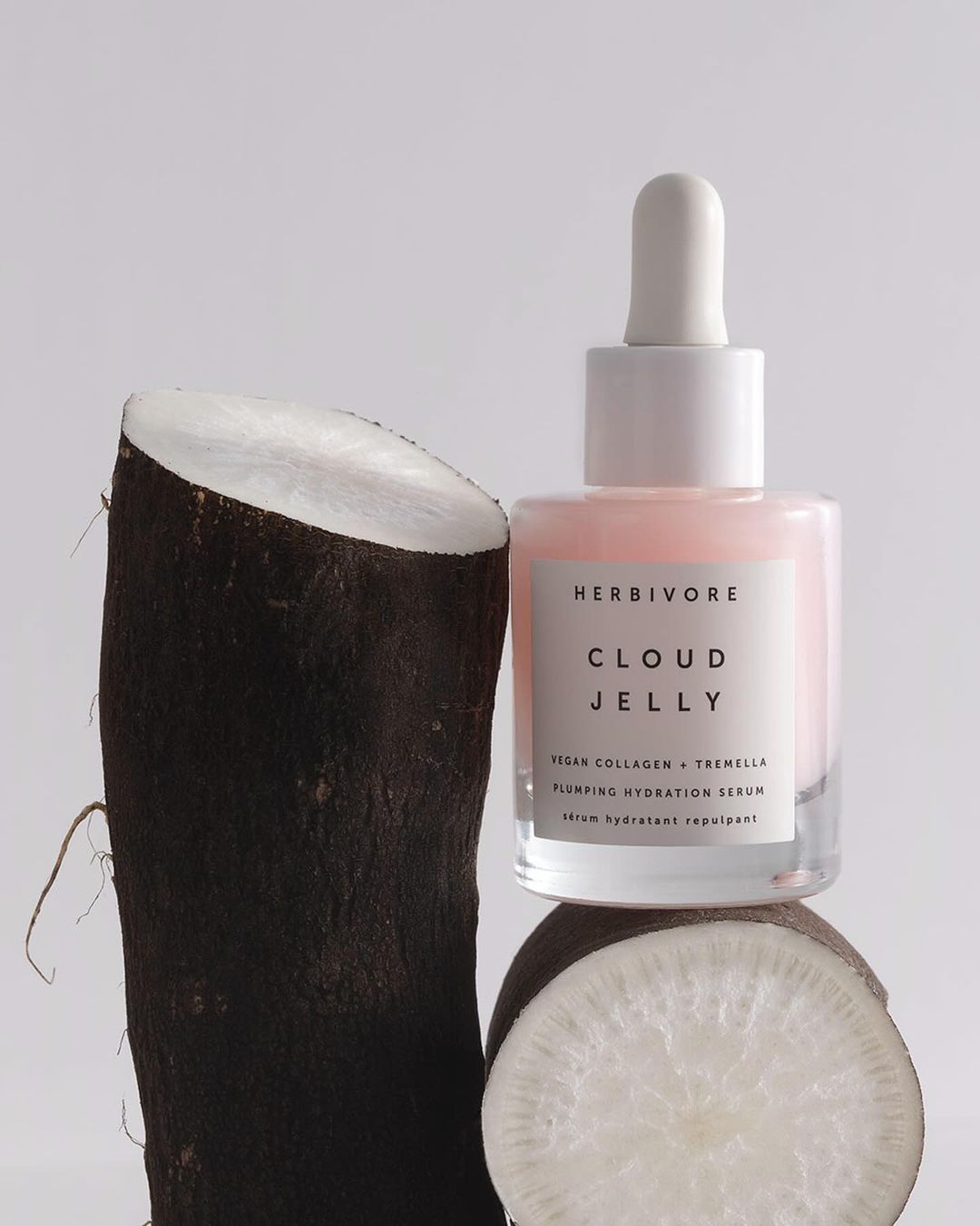 Close-up studio shot of Herbivore Cloud Jelly serum bottle alongside cut yam root against a white background, product photography.