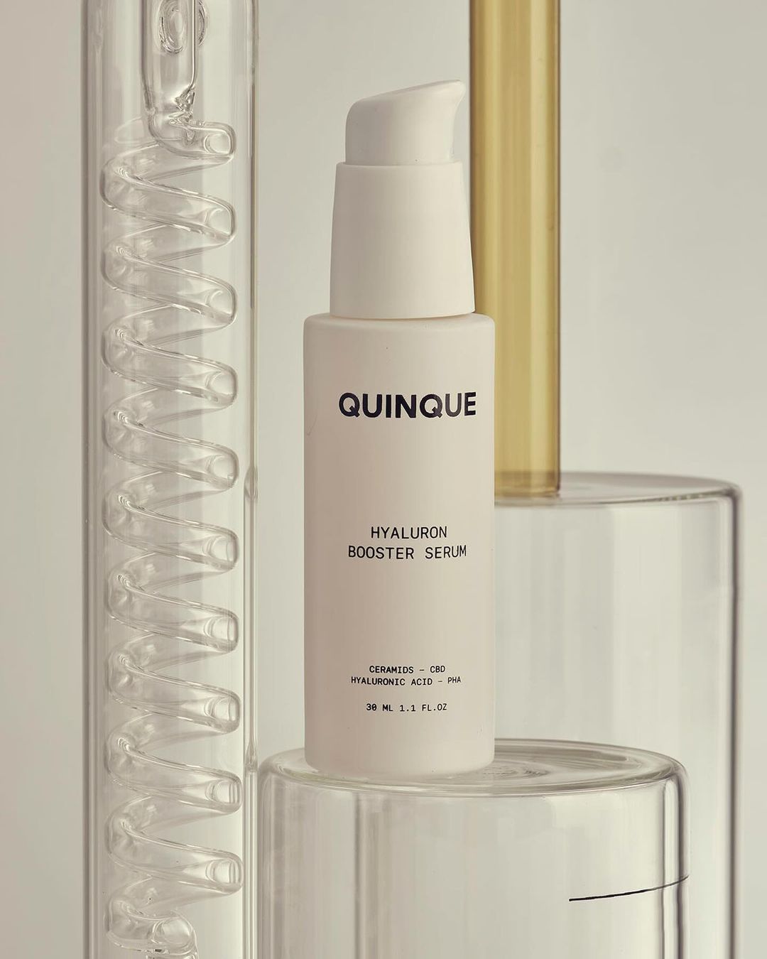 Close-up of QUINQUE Hyaluron Booster Serum bottle with pump on clear geometric stands, against a soft white background.