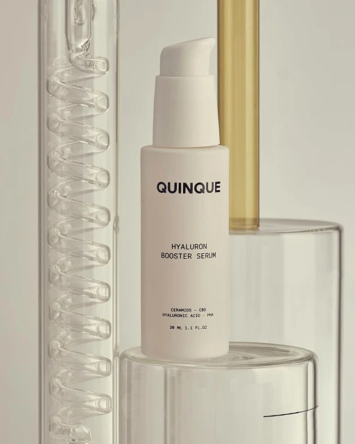Close-up of QUINQUE Hyaluron Booster Serum bottle with pump on clear geometric stands, against a soft white background.