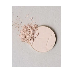 Round pressed powder compact with loose powder scattered on a textured light grey surface by photographer Alice Cuvelier.