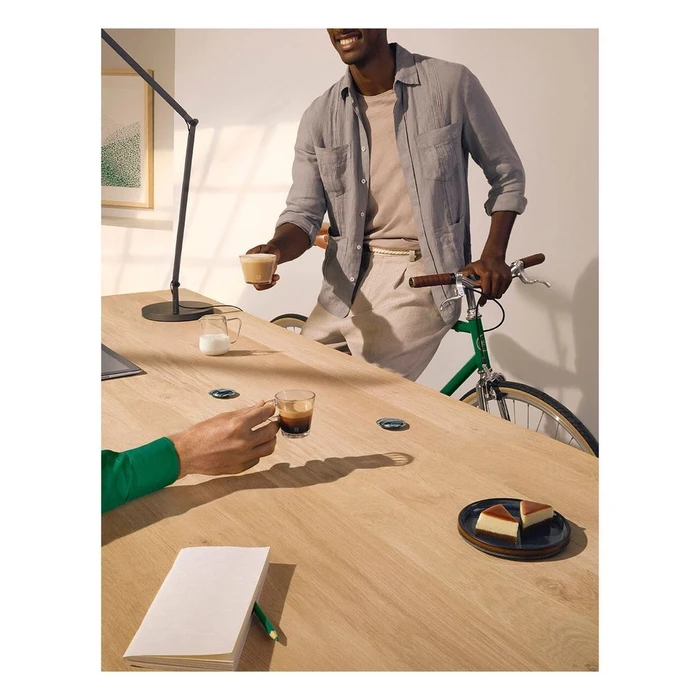 Man holding a small coffee cup standing next to a green bicycle beside a long wooden table with pastry and espresso.