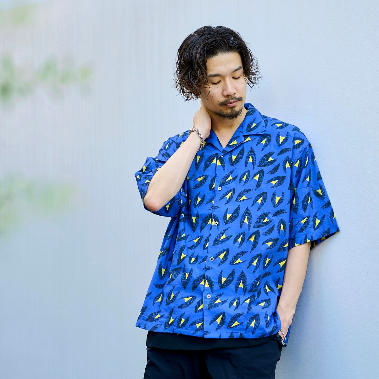 Adult male in casual patterned shirt poses outdoors, looking down. He has dark hair and a beard, standing against a muted background.