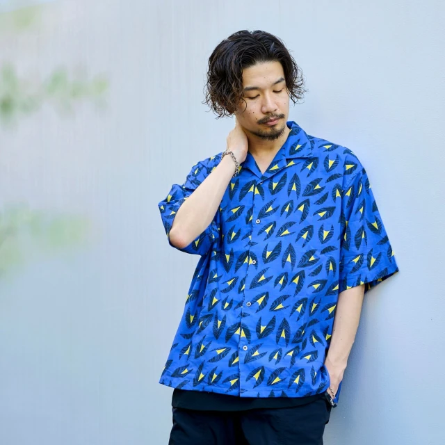 Adult male in casual patterned shirt poses outdoors, looking down. He has dark hair and a beard, standing against a muted background.