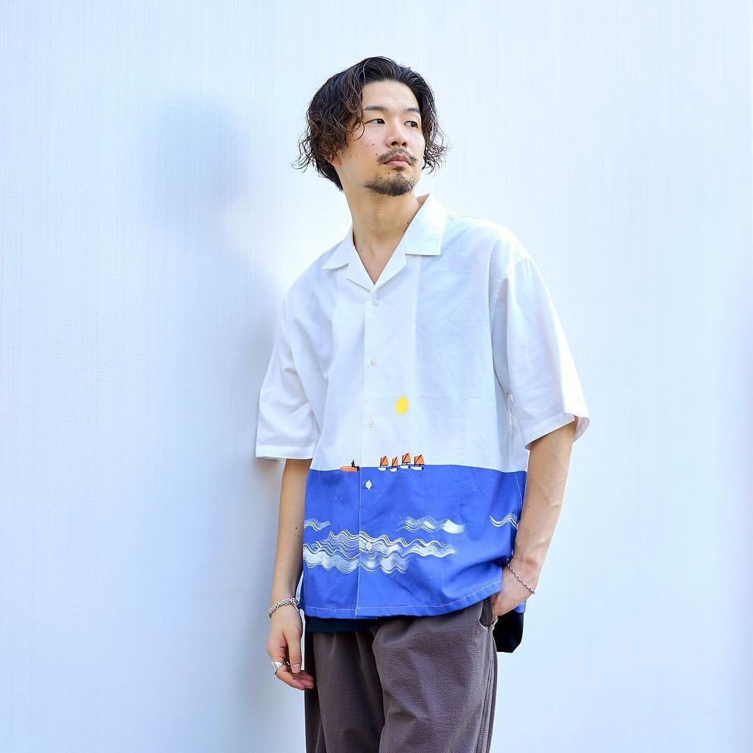 Adult male in a button-down shirt with ocean-themed print, stands against a white wall with curly hair and a subtle mustache.