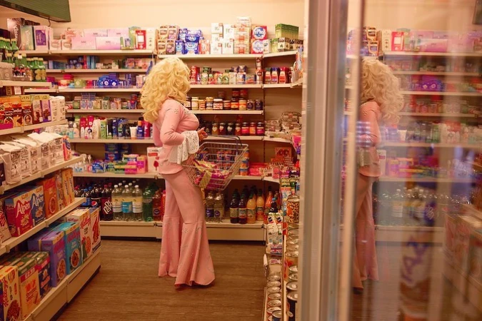 Woman in pink outfit and blonde wig shops in grocery store. Shelves filled with various food items provide background in indoor setting.