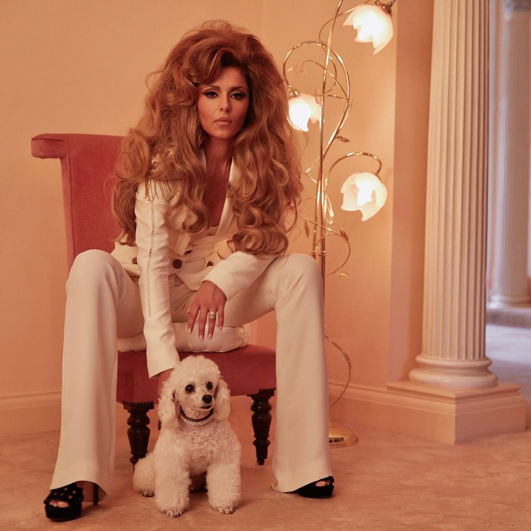Woman in white suit poses with a white poodle indoors with a decorative lamp and classic architecture in the background.