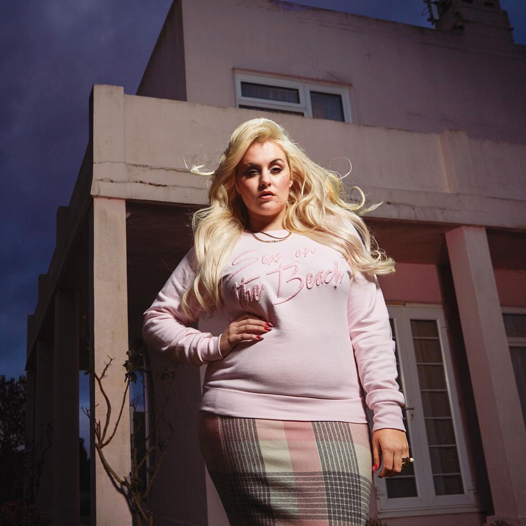 Portrait of an adult female with long blonde hair, wearing a pink sweatshirt and plaid skirt, posing in front of a pink building.