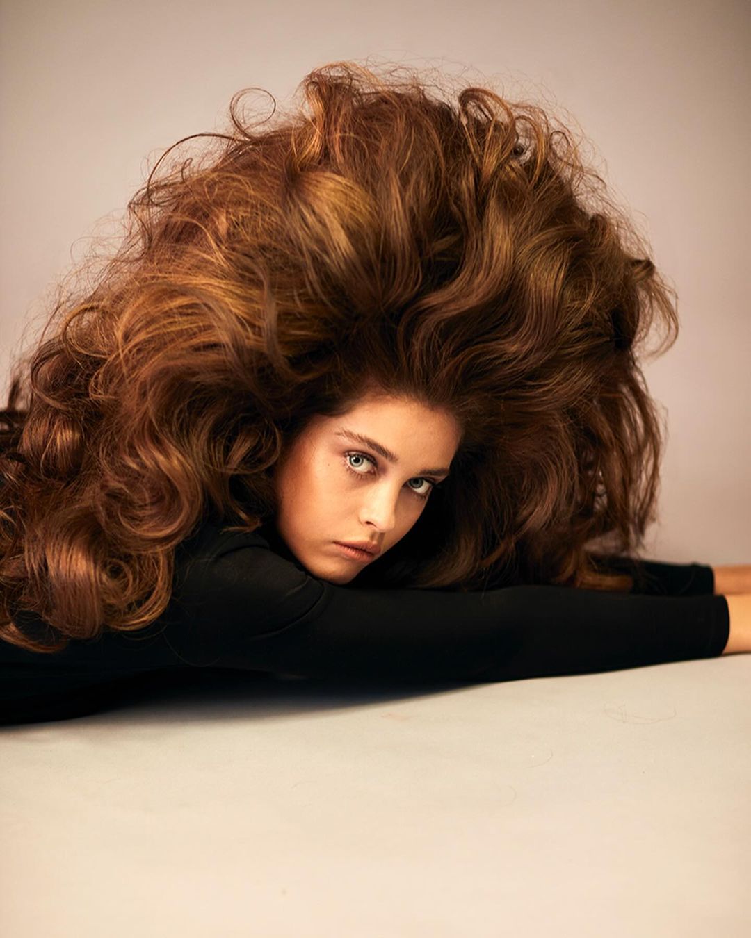 Portrait of a young woman with light eyes and voluminous brown hair, lying down in a studio with a soft, neutral background.