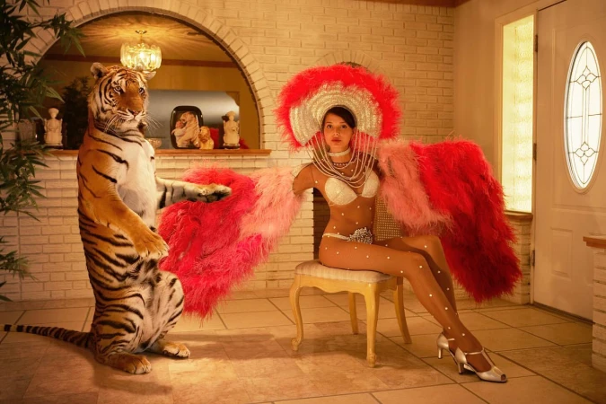 A human and a tiger stand together indoors, the woman wearing a feathered costume while seated on a chair, under warm indoor lighting.