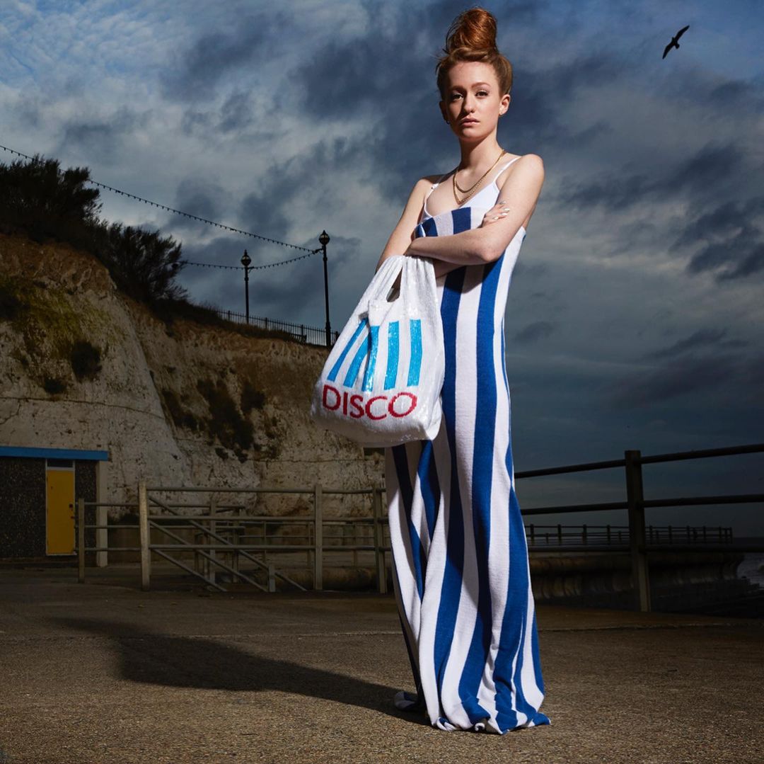 A slender woman in a striped dress stands on a seaside promenade holding a 'DISCO' bag with a cloudy sky and cliffs behind her.