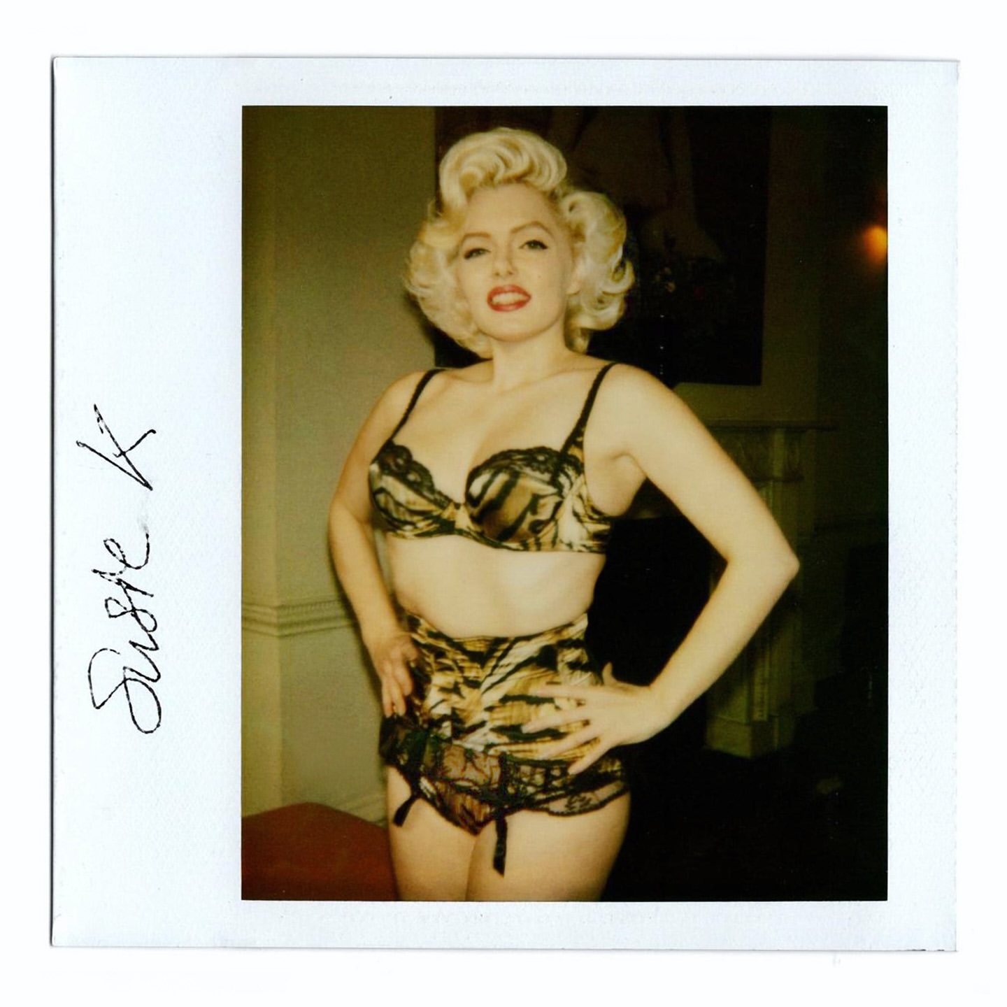Portrait of an adult female with blonde curled hair posing indoors wearing a tiger print lingerie set against a neutral background.