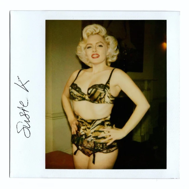 Portrait of an adult female with blonde curled hair posing indoors wearing a tiger print lingerie set against a neutral background.