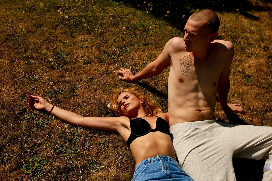 A shirtless man sits beside a blonde woman in jeans and a bra top, relaxing on grass in sunny daylight in an outdoor setting.