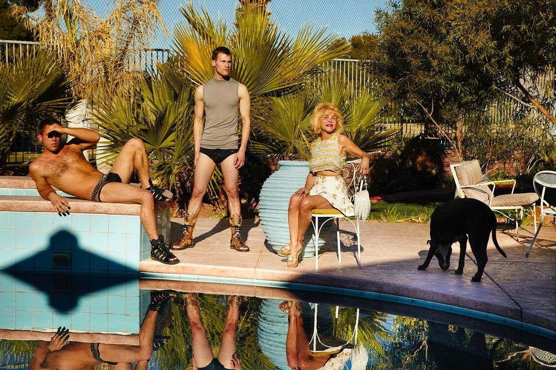 Three people and a black dog pose beside a pool outdoors, with palm trees and landscaping in the background on a sunny day.