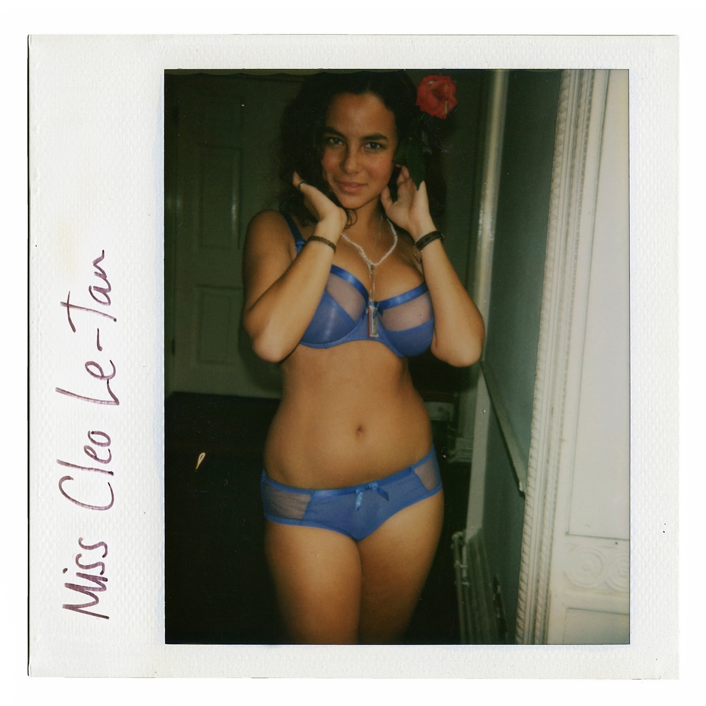Young woman in matching blue lingerie stands indoors, framed by a vintage polaroid border. A red flower is tucked behind her ear.