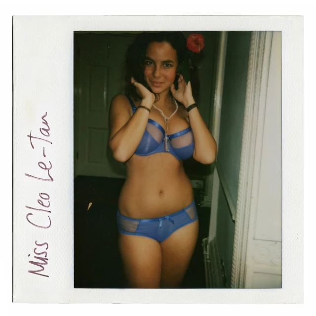 Young woman in matching blue lingerie stands indoors, framed by a vintage polaroid border. A red flower is tucked behind her ear.