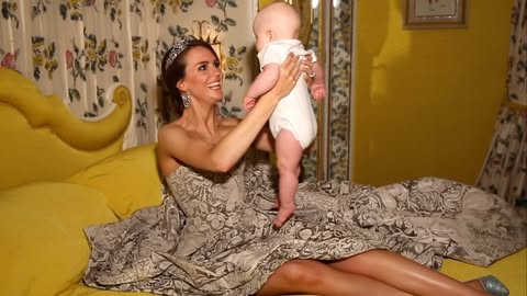 Woman in strapless dress and tiara sits on a yellow bed, holding a baby wearing white onesie in a bright yellow room with floral curtains.