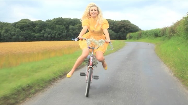 Adult female riding a bicycle on a country road. She wears a short yellow dress and matching flats, appears happy and is jumping.