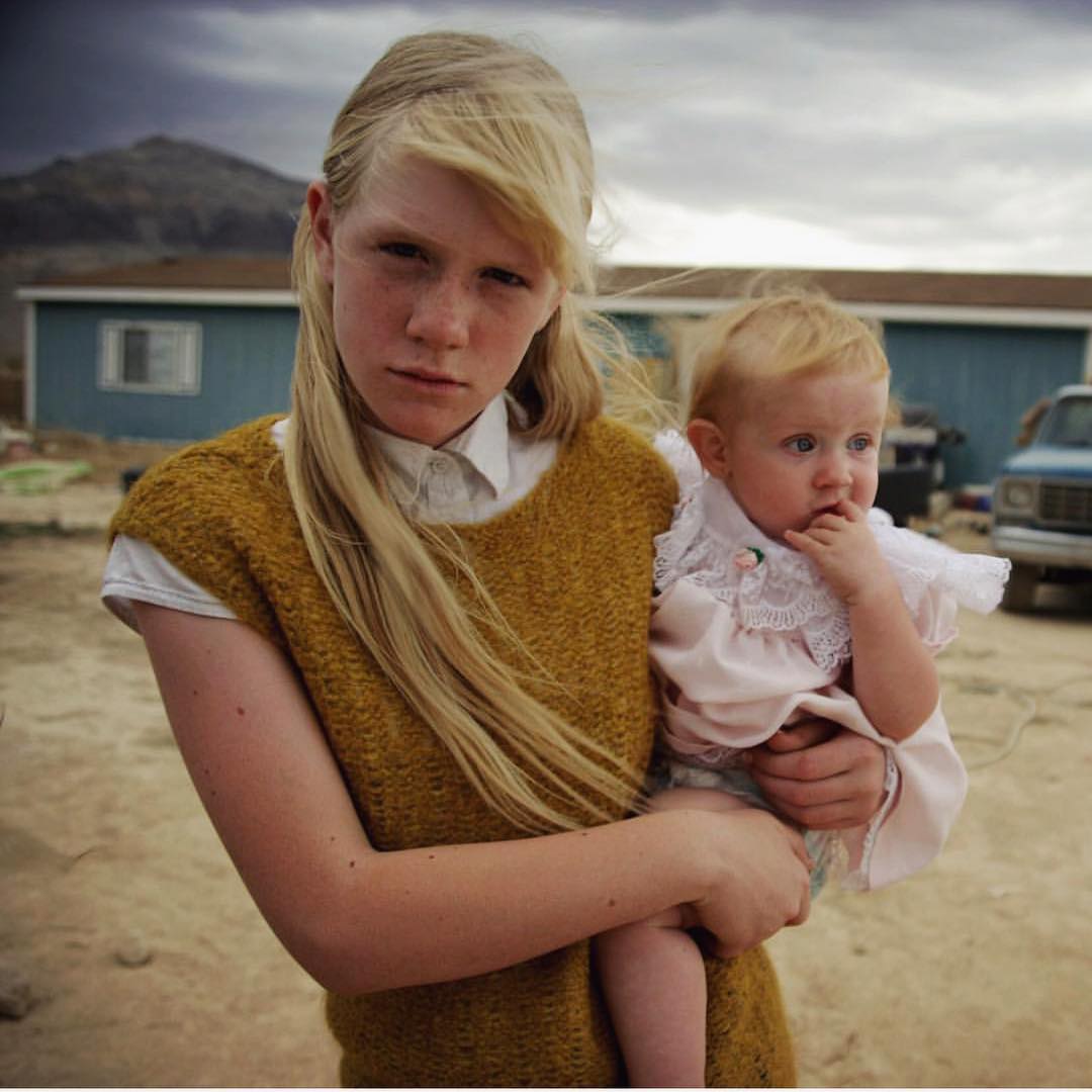 Portrait of a young adult female holding a baby outdoors in a dry landscape near a blue house, beneath a cloudy sky with distant mountains.