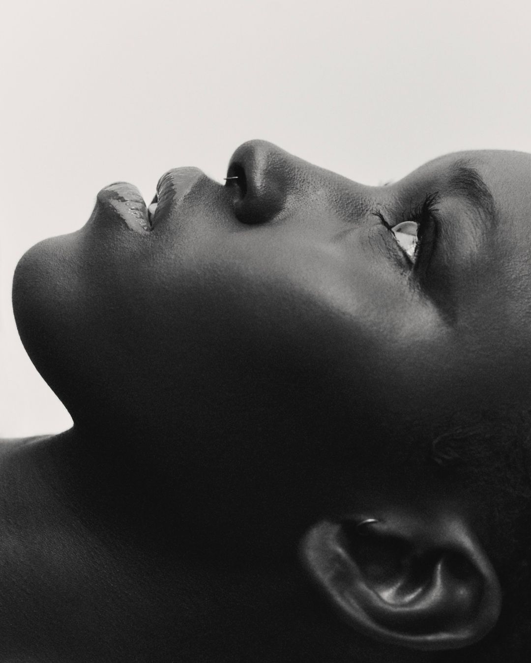 Monochromatic close-up portrait of a Black adult looking upwards, featuring a minimalist studio background, highlighting skin texture and tone.