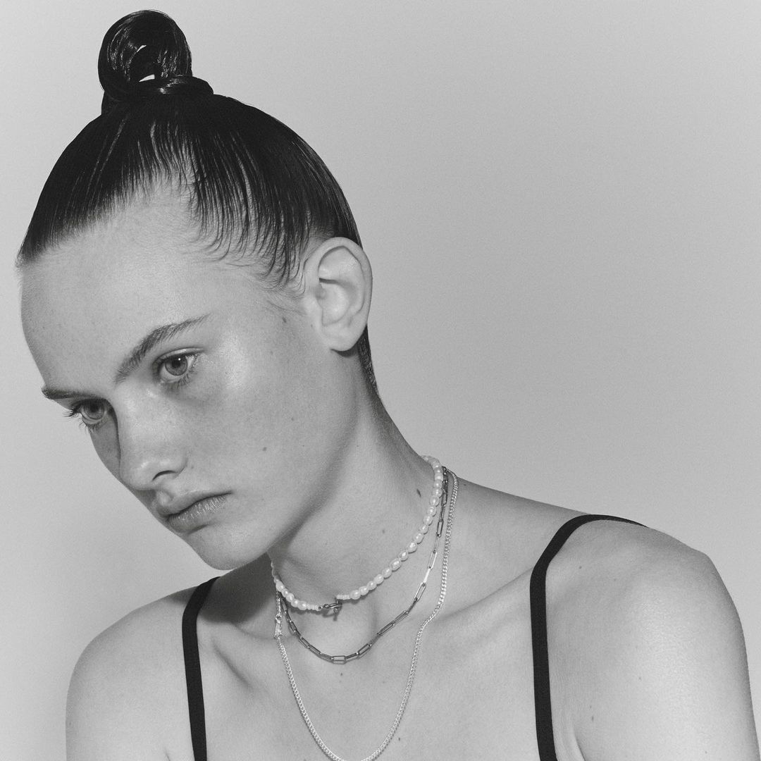 Close-up black and white portrait of a young adult woman with a sleek bun wearing multiple layered necklaces against a white backdrop.