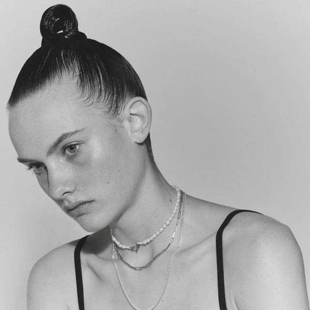 Close-up black and white portrait of a young adult woman with a sleek bun wearing multiple layered necklaces against a white backdrop.