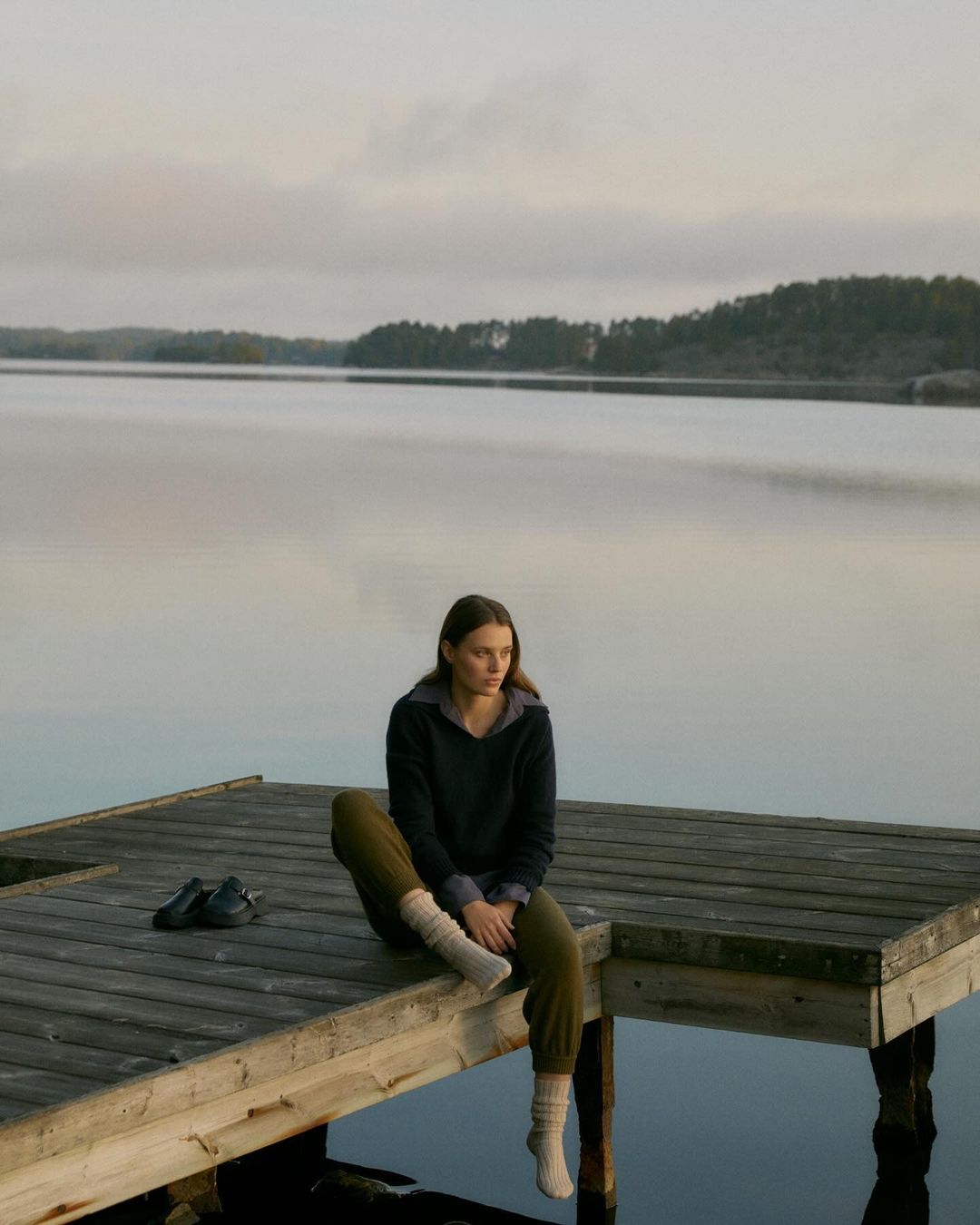 A young woman in casual clothing sits on a wooden dock with her legs dangling, overlooking a tranquil lake and forested shoreline under cloudy skies.