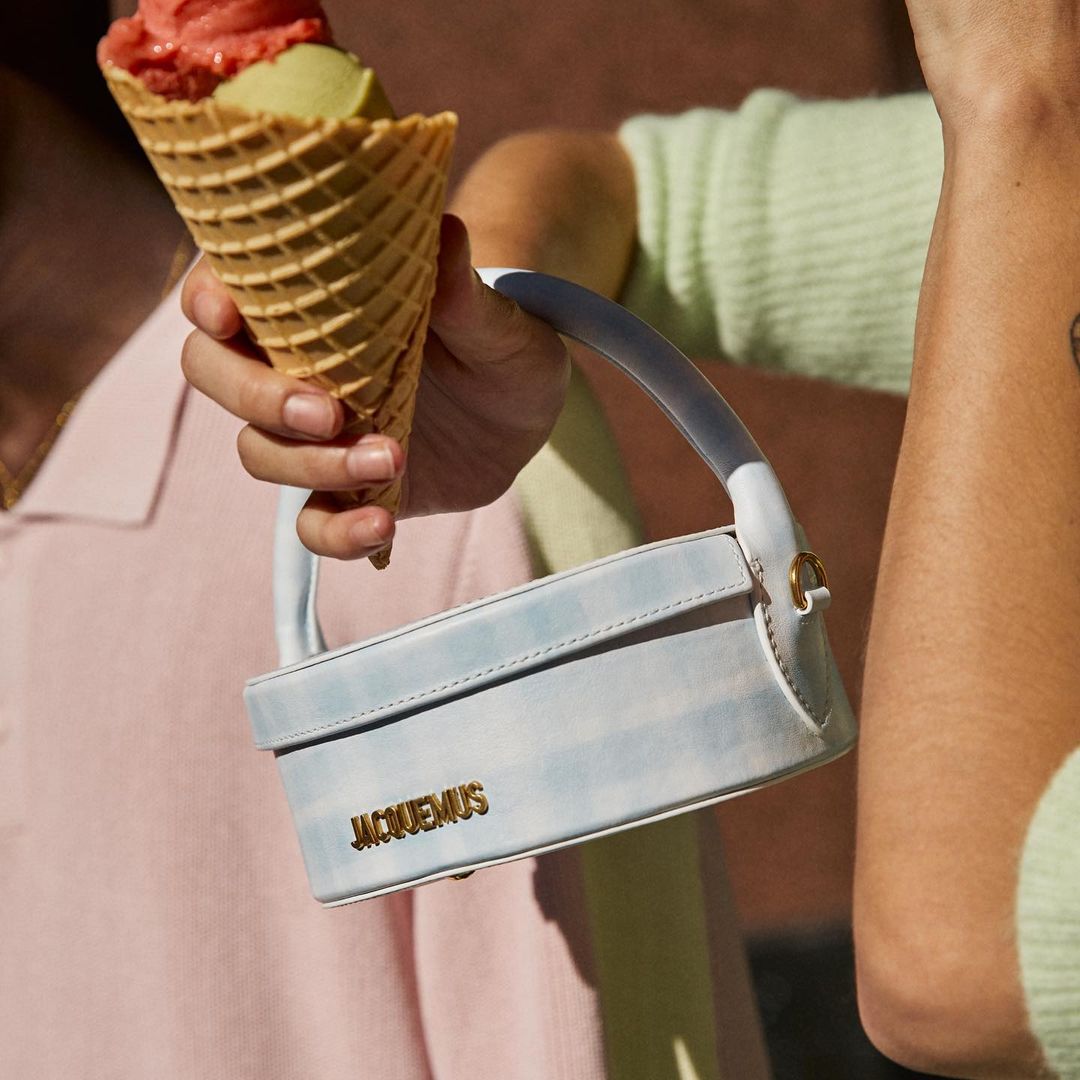 Woman holds an ice cream cone and Jacquemus handbag. The cone features red and green ice cream scoops, set against a light brown backdrop.