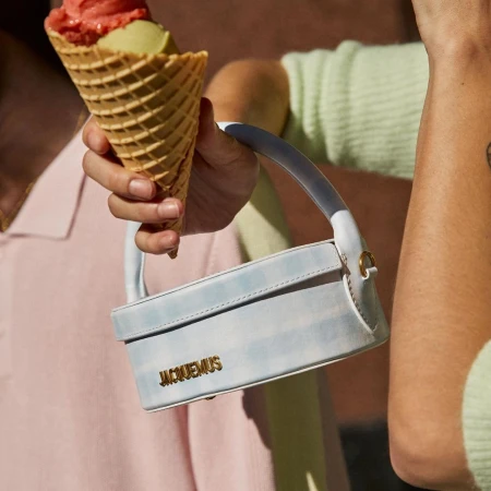 Woman holds an ice cream cone and Jacquemus handbag. The cone features red and green ice cream scoops, set against a light brown backdrop.